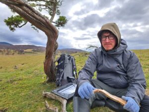 A man sitting in front of a tree with a rucksack and survey notepad, facing the camera with an upland landscape behind