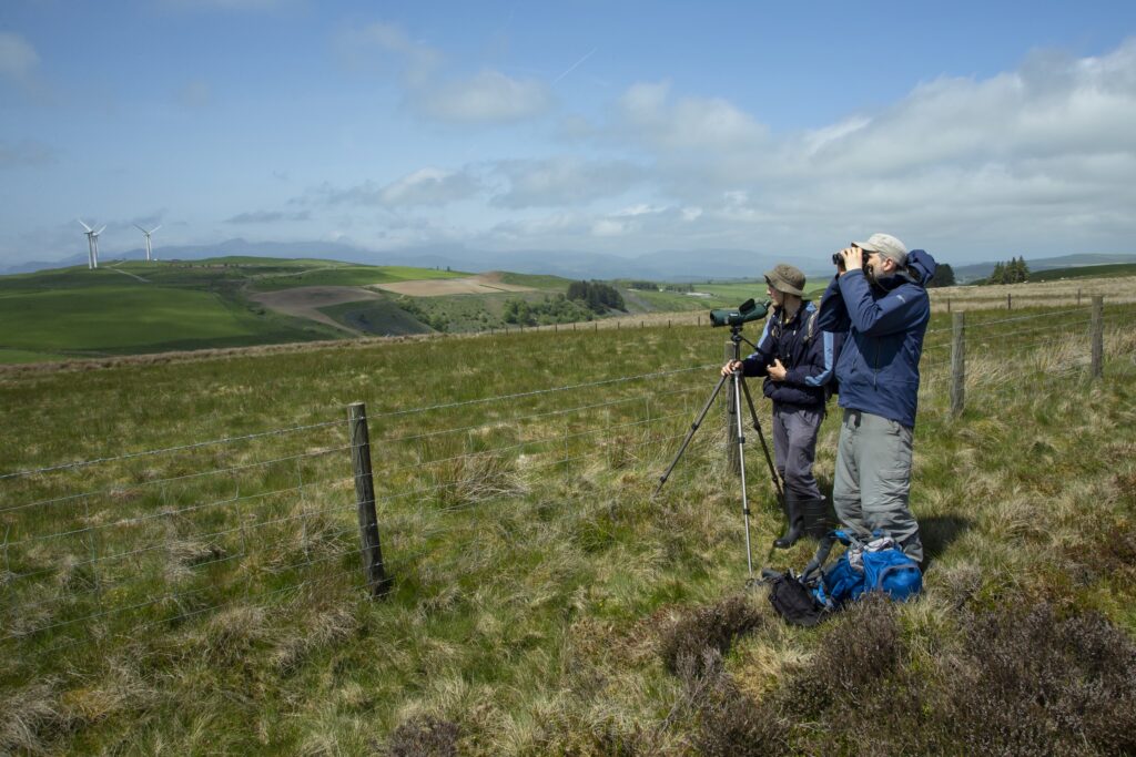 A man with binoculars and another with a spotting scope looking out across an upland field.